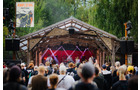 Download Foto: Open-Air-Bühne mit Band, umgeben von Bäumen. Viele Zuschauer im Vordergrund. Stimmung fröhlich und entspannt.
