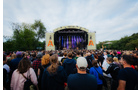 Download Foto: Open-Air-Konzert bei Tag: Menschenmenge steht vor einer Bühne mit Lichtern im Freien, Bäume im Hintergrund, bewölkter Himmel.