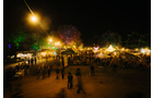 Download Foto: Nachtszene eines lebhaften Festes im Freien mit vielen beleuchteten Ständen und Besuchern, Bäume im Hintergrund sind bunt erleuchtet.