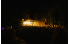 Download Foto: Eine Bühne bei Nacht, beleuchtet von gelbem Licht, mit Menschenmenge im Vordergrund und Bäumen im Hintergrund.