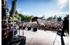 Download Foto: Eine Bühne im Freien mit Musiker beim Sprung in die Luft, vor einem großen, begeisterten Publikum unter strahlendem Himmel.