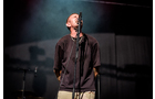 Download Foto: Eine Person steht singend auf einer Bühne vor einem Mikrofon, mit dunkler Kleidung und beleuchtet von Bühnenlicht.