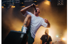 Download Foto: Ein Sänger in weißem T-Shirt und grüner Hose performt energiegeladen auf einer Bühne, Mikrofon in der Hand, Bühnenscheinwerfer leuchten.