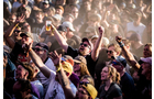 Download Foto: Menschenmenge auf einem Festival, fröhlich tanzend und feiernd. Einige heben die Hände, einer hält ein Bier. Staubig und lebhaft.