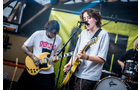 Download Foto: Zwei Musiker spielen E-Gitarren auf einer Bühne, einer singt ins Mikrofon. Im Hintergrund ist ein Schlagzeug zu sehen.