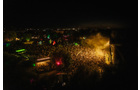 Download Foto: Luftaufnahme eines Festivals bei Nacht: Menschenmenge vor Bühne mit farbigen Lichtern und Rauch. Dunkler Himmel im Hintergrund.