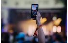Download Foto: Eine Person hält ein Smartphone hoch, um eine Konzertbühne im Hintergrund zu filmen. Verschwommene Lichter und Menschenmenge sind sichtbar.