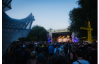 Download Foto: Open-Air-Konzert bei Dämmerung mit einer Menschenmenge vor einer beleuchteten Bühne, umgeben von Bäumen und einem Zeltdach.