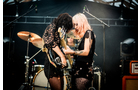 Download Foto: Zwei Musikerinnen spielen E-Gitarre auf einer Bühne. Sie tragen glitzernde Outfits, im Hintergrund sind Schlagzeug und Verstärker.
