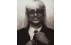 Download Foto: Schwarz-weiß Foto einer Person mit langen, platinblonden Haaren und Brille, die einen Anzug trägt und ihre Krawatte richtet.