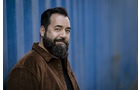 Download Foto: Ein Mann mit Bart und brauner Jacke steht vor einer blauen Wand und lächelt leicht in die Kamera.