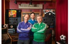 Download Foto: Drei Männer in bunten Trainingsjacken stehen in einem Tonstudio, umgeben von Lautsprechern und Fußbällen.