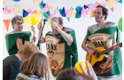 Download Foto: Drei Personen in Kostümen mit 'Baked Beans'-Aufdruck musizieren fröhlich vor Publikum. Bunte Girlanden schmücken den Hintergrund.