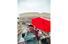 Download Foto: Ein lächelnder Mann mit rotem Regenschirm steht vor dem Kolosseum. Es regnet leicht, die Straße ist belebt.