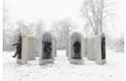 Download Foto: Sechs schneebedeckte Steinskulpturen mit menschlichen Figuren stehen in einer verschneiten Landschaft, umgeben von kahlen Bäumen.
