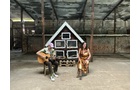 Download Foto: Zwei Personen in einer Lagerhalle, eine spielt Gitarre, die andere sitzt daneben. Im Hintergrund ein kleines, dreieckiges Häuschen.