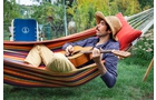 Download Foto: Mann mit Strohhut liegt in bunter Hängematte im Garten und spielt Gitarre, umgeben von grünem Rasen und Pflanzen.