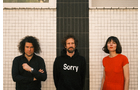 Download Foto: Drei Personen stehen vor einer gefliesten Wand. Die Person in der Mitte trägt einen Pullover mit der Aufschrift 'Sorry'.