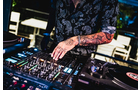 Download Foto: Ein DJ mit tätowiertem Arm bedient ein Mischpult. Im Hintergrund sind verschwommene Lichter und Möbel erkennbar.