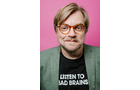 Download Foto: Ein lächelnder Mann mit Brille, grünem Sakko und T-Shirt mit der Aufschrift 'LISTEN TO BAD BRAINS' vor einem pinken Hintergrund.