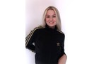 Download Foto: Eine lächelnde Person mit langen blonden Haaren trägt einen schwarzen Adidas-Trainingsanzug mit goldenen Streifen vor weißem Hintergrund.