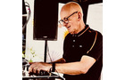 Download Foto: Ein lächelnder älterer Mann mit Brille und schwarzem Hemd steht an einem DJ-Pult und bedient die Geräte.