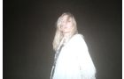Download Foto: Eine Person mit langem, blondem Haar trägt ein weißes Outfit und schaut in die Kamera, vor einem dunklen, nebligen Hintergrund.