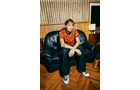 Download Foto: Eine Person sitzt in einem Aufnahmestudio auf einem schwarzen Ledersofa. Sie trägt ein orangefarbenes Shirt und schwarze Hosen.