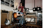 Download Foto: Zwei Personen in einem Tonstudio, eine sitzt mit Gitarre, die andere steht hinter einem Mischpult, umgeben von Lautsprechern.