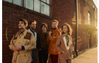 Download Foto: Fünf Personen in Retro-Outfits stehen nebeneinander vor einer Backsteinmauer, beleuchtet von warmem Sonnenuntergangslicht.