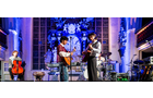 Download Foto: Eine Band spielt auf einer beleuchteten Bühne in einer großen Halle, umgeben von Instrumenten und einer beeindruckenden Wanddekoration.