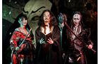 Download Foto: Vier Personen in aufwendigen, dunklen Kostümen und Schminke stehen vor einer dramatischen Kulisse, die an ein Theater erinnert.