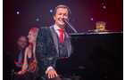 Download Foto: Ein Mann in glitzerndem Anzug singt fröhlich am Klavier, auf dem ein Getränk steht. Hintergrund unscharf, rote Bühnenbeleuchtung.