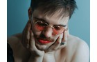 Download Foto: Eine Person mit roten Lippen und rosa Brille schaut nachdenklich. Hände stützen das Gesicht, Hintergrund ist blau.