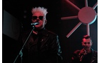 Download Foto: Ein Sänger mit weißem Haar und Sonnenbrille singt in ein Mikrofon. Ein Musiker im Hintergrund spielt Gitarre, beleuchtet von rotem Licht.