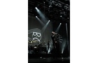 Download Foto: Ein Sänger tritt auf einer Bühne mit dunkler Beleuchtung auf. Im Hintergrund sind Bandmitglieder und ein großes Logo zu sehen.