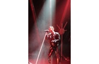 Download Foto: Eine Person singt mit einem Mikrofon auf einer Bühne, umgeben von rotem Licht und Rauch, trägt ein auffälliges, glänzendes Outfit.