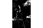 Download Foto: Schwarz-weiß-Bild einer Person mit Lederjacke, die ein Mikrofon hält, vor einer Bühne mit Instrumenten.