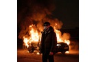 Download Foto: Person steht nachts vor brennendem Auto, trägt Mantel und Mütze. Flammen und Rauch steigen in den dunklen Himmel auf.