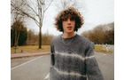 Download Foto: Junger Mann mit lockigem Haar und gestreiftem Pullover steht auf einer Straße, umgeben von kahlen Bäumen an einem bewölkten Tag.