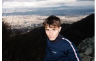 Download Foto: Ein junger Mann steht auf einem Hügel mit Blick auf eine Stadtlandschaft im Hintergrund, bei bewölktem Himmel.