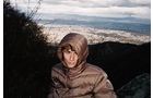 Download Foto: Eine Person in einem braunen Mantel steht vor einer Landschaft mit Bergen und einer Stadt im Hintergrund. Der Himmel ist wolkig.