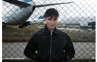 Download Foto: Ein junger Mann steht vor einem Maschendrahtzaun auf einem Flugfeld. Im Hintergrund sind Flugzeuge zu sehen.