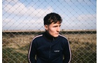 Download Foto: Ein junger Mann steht vor einem Maschendrahtzaun auf einem Feld, trägt eine dunkelblaue Jacke und schaut nach rechts.