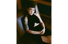 Download Foto: Person mit kurzem Haar sitzt auf einem Stuhl, trägt ein ärmelloses Kleid und hat ein Tattoo am Arm. Stimmungsvolle Beleuchtung.