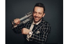 Download Foto: Ein lächelnder Mann in kariertem Anzug hält eine Trophäe und zeigt seine Armbanduhr vor einem dunklen Hintergrund.
