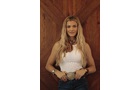 Download Foto: Eine Person mit langen, blonden Haaren trägt ein weißes Oberteil und Jeans. Sie steht vor einem hölzernen Hintergrund.