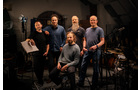 Download Foto: Fünf Männer in einem Musikstudio, umgeben von Instrumenten. Vier stehen, einer sitzt. Sie lächeln und wirken kameradschaftlich.