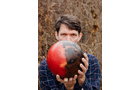 Download Foto: Ein Mann mit kariertem Hemd hält einen rot-schwarzen Ball, der sein Gesicht verdeckt, vor einem Hintergrund aus trockenem Laub.
