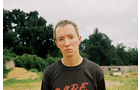 Download Foto: Junger Mensch steht draußen vor bewaldetem Hintergrund, blickt ernst in die Kamera, trägt ein dunkles T-Shirt.